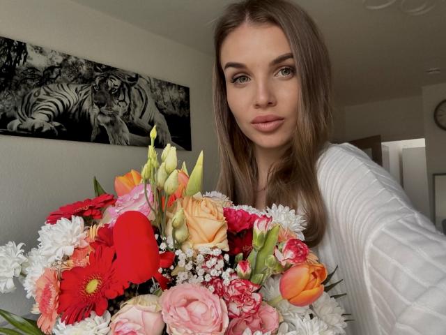 LightTeresa met bruin haar, gekleed in een witte geribbelde top, die een kleurrijke bos rozen, roze bloemen en rode tulpen vasthoudt, en die in de camera kijkt met een lichte glimlach..
