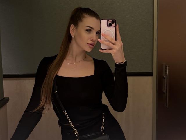 LightTeresa con una lunga coda di capelli castani, una maglia nera a maniche lunghe, pantaloni neri, che tiene il telefono allo specchio, mostrando il suo seno piccolo e il sedere slanciato, in piedi in una stanza poco illuminata..