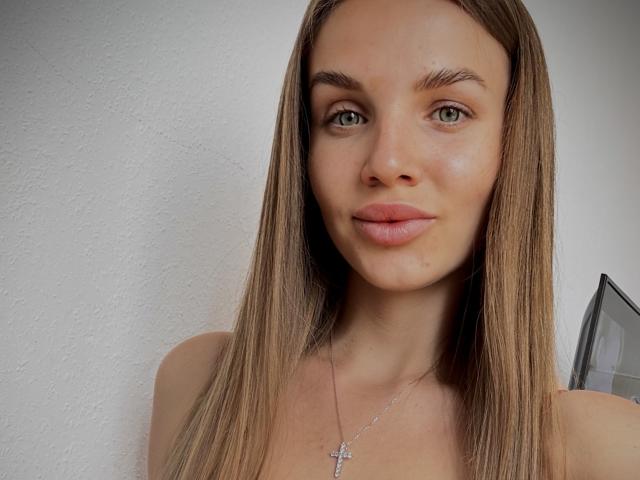 LightTeresa has long, straight brown hair, green eyes, and pink lips, wearing a silver cross necklace. She's smiling subtly with a slight tan and a clean, white wall behind.