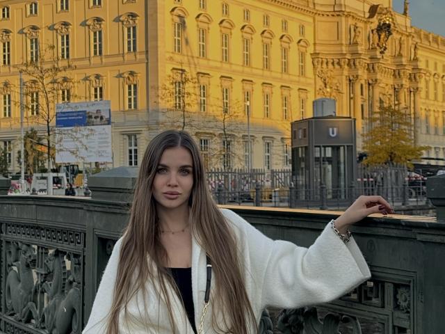 LightTeresa, avec de longs cheveux bruns, un manteau blanc, un haut noir, debout près d'une balustrade en fer vert, le bras droit reposant, regardant la caméra, un bâtiment jaune en arrière-plan, lumière du soir..