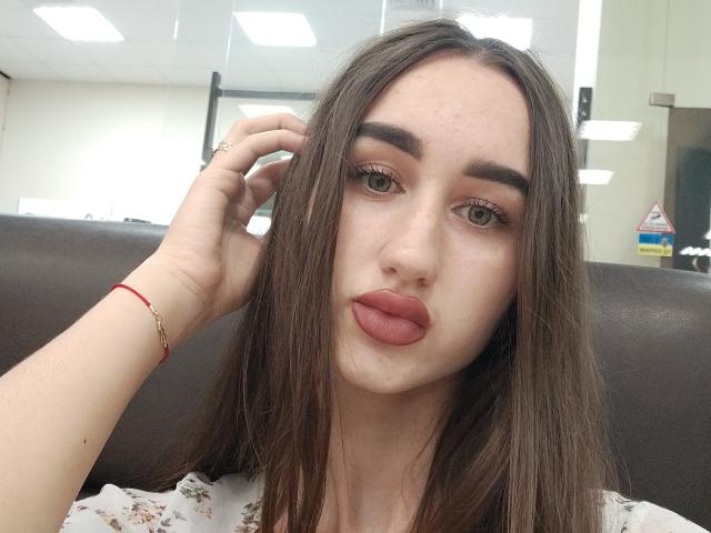 FortuneX with long brown hair, pouty pink lips, and green eyes, looking at the camera, hand in hair, wearing a red bracelet, in a bright indoor setting.