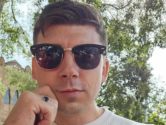 AlexxStar with short brown hair, wearing black reflective sunglasses, white shirt, and black ring on right hand, resting chin on hand, looking serious. Sunlight through green tree leaves.