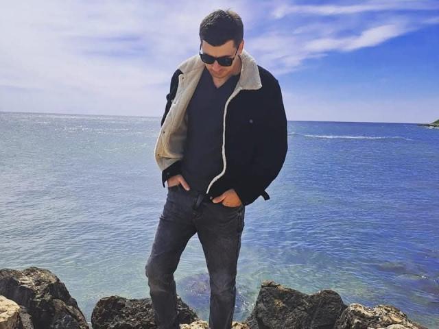 AlexxStar stands by rocky shore, wearing black jacket with beige lining, dark sunglasses, gray pants, hands in pockets, looking down, with clear blue ocean and sky in background.