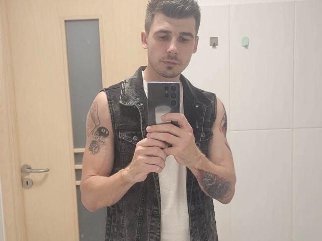 AlexxStar with tattoos on arms, wearing a black sleeveless denim vest and white tee, standing in a bathroom, taking a mirror selfie with a smartphone.