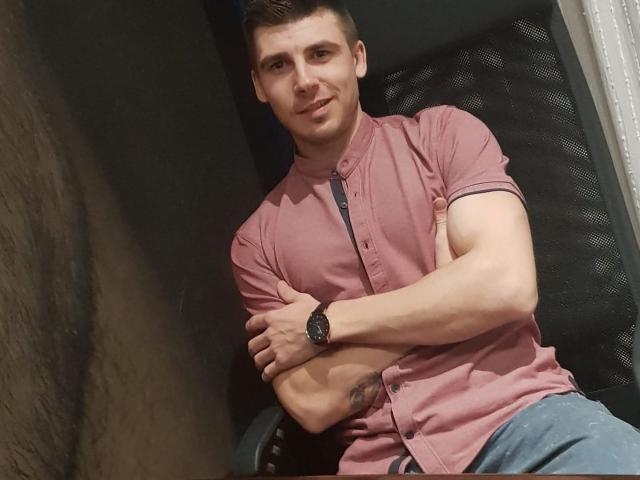 AlexxStar, muscular, wearing pink button-up shirt, crossed arms, blue jeans, black watch, short brown hair, slight smile, tattoo on right forearm, standing in dimly lit room.