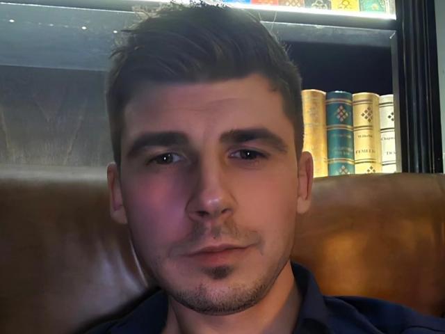 AlexxStar has a handsome face with short brown hair, light skin, and a small goatee. He's wearing a dark shirt, sitting in a brown leather booth.