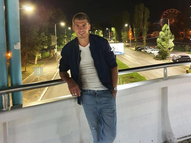 AlexxStar, standing with hands in pockets, wearing a blue shirt over a white tee, and blue jeans, leaning on a railing at night, smiling. Nightlight glow.
