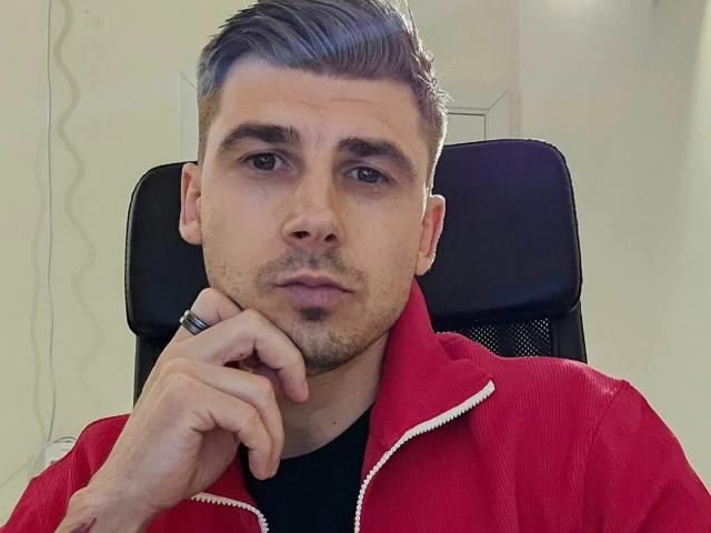 AlexxStar with short, slicked-back gray hair, wearing a red zip-up jacket, black shirt, and a ring on his right hand, looking serious, resting his chin on his hand.