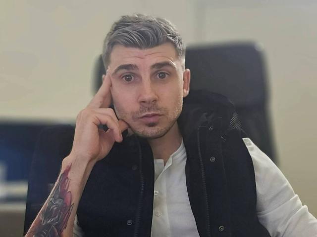 AlexxStar with gray-styled hair, serious expression, white shirt, black vest, tattooed arm, hand on cheek, in office chair.