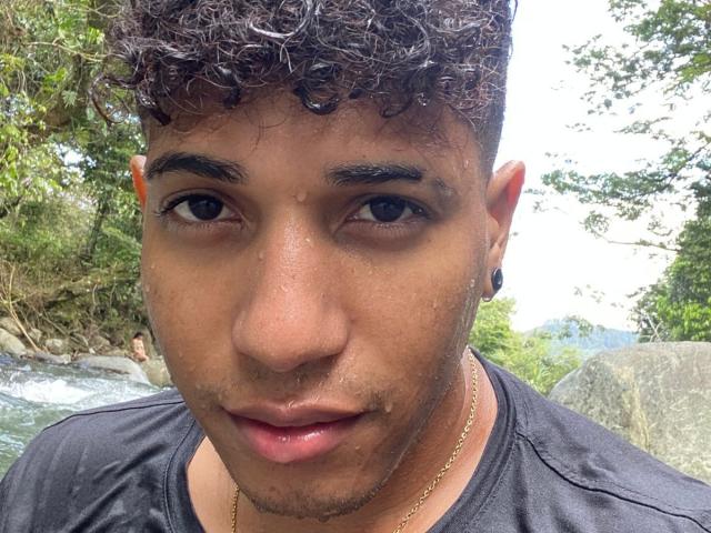Yoximanuelprime tiene el pelo castaño rizado, piel oscura y una cadena de oro. Lleva una camisa negra, con un piercing en la nariz y aretes grandes. Está mirando a la cámara..