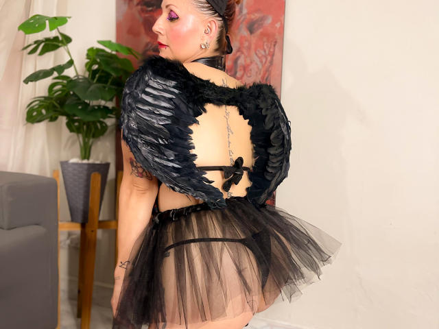GiselleDunn wearing black feathered wings, sheer black skirt, black bra, and thong, showing her ass, with a chain on her back, and pink eye makeup.