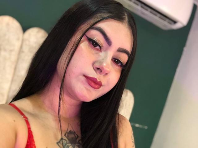AlanaaWalker has long black hair, red lipstick, and thick eyebrows. She's wearing a red top, showing a chest tattoo, and has a slight smile. Her makeup is bold.
