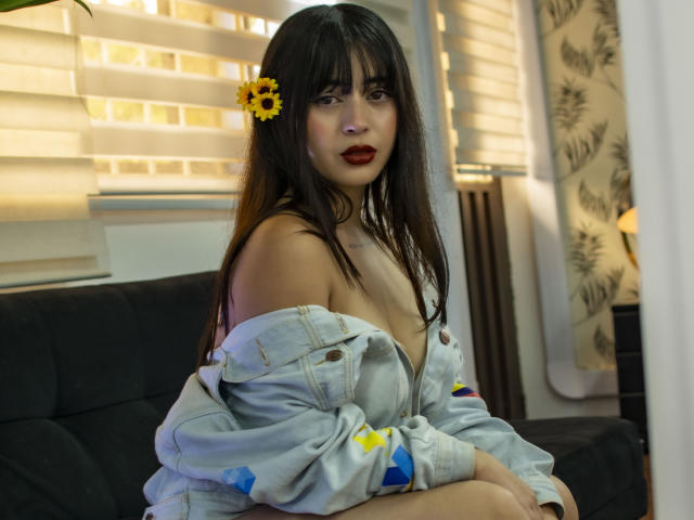 SopphieBloom with sunflower hair clip, dark lipstick, off-shoulder denim jacket, showing tits, sitting on black couch, looking at camera, sunlight through blinds, soft brown eyes.