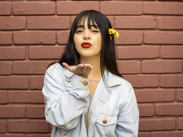 SopphieBloom with dark hair, yellow flower, red lipstick, and denim jacket, making a "shushing" gesture, showing off her tits slightly, against a brick wall.