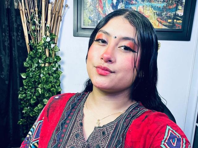 KalpanaAisha tiene el cabello oscuro y húmedo, maquillaje rojo en los ojos y un pequeño punto en la frente. Lleva una blusa roja con estampado y un collar, y frunce ligeramente los labios..