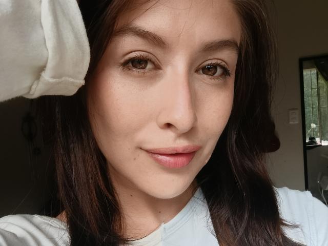 IsabellaPalacio has brown eyes, light skin, and brown wavy hair. She's wearing a white shirt, smiling slightly, with a white towel on her head. Her lips are pink.