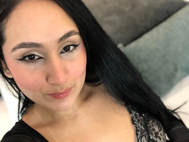 HiilaryDolce has black hair, brown eyes with winged eyeliner, pink lips, and a light skin tone. She's wearing a black lace top, looking at the camera.