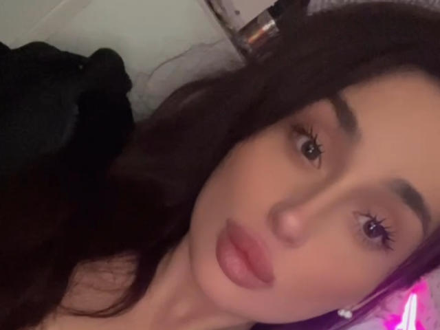 MollyyDi has dark brown hair, full eyebrows, and pink lips. Her eyes are half-closed, and she's looking slightly to the side. She's wearing a black top.