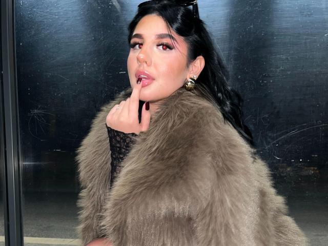 Carolineyourfav-hot has long black hair, wearing a brown fur coat. She's looking over her shoulder with a finger to her lips, slightly parted pink lips. She's got gold hoop earrings and a black lace sleeve. Her makeup is bold with red eyeliner. The fur coat is textured and fluffy. She's in front of a glass window with some smudges. The light is soft, highlighting her face and fur coat. The overall vibe is chic and slightly mysterious.