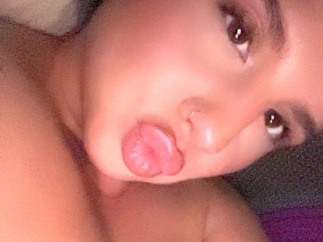 Carolineyourfav-hot's face, tongue out, pink lips, brown eyes, small nose, slightly blurred, with a visible nose ring, in a selfie, upper chest visible, no clothes shown.