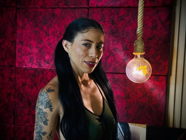 KattySloven mit schwarzen Zöpfen, einem grünen Tanktop, einem tätowierten Arm und rotem Lippenstift, lächelt zu einer hängenden Glühbirne, wobei ihre Brüste leicht sichtbar sind, in einem schwach beleuchteten Raum..