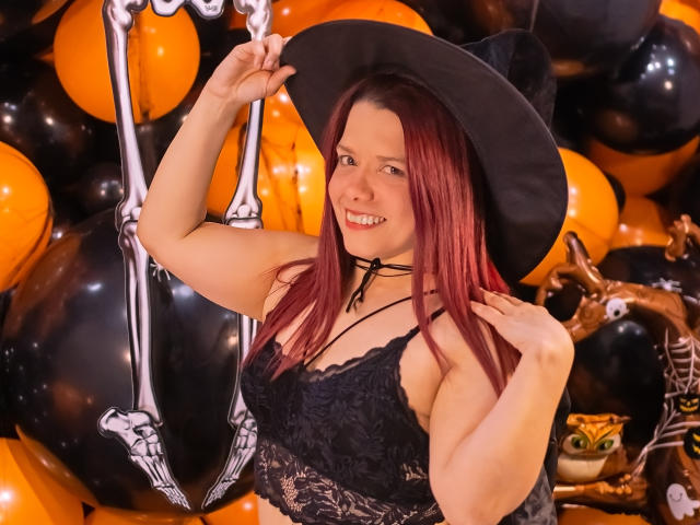 BiancaWild with red hair, black lace top, showing her tits, wearing a black witch hat, smiling, holding a skeletal balloon, surrounded by orange and black balloons.