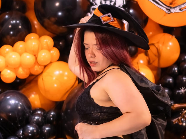 BiancaWild with red hair, wearing a black lace top and witch hat, holding her tits, surrounded by orange and black balloons. She's looking down, slightly biting her lip.