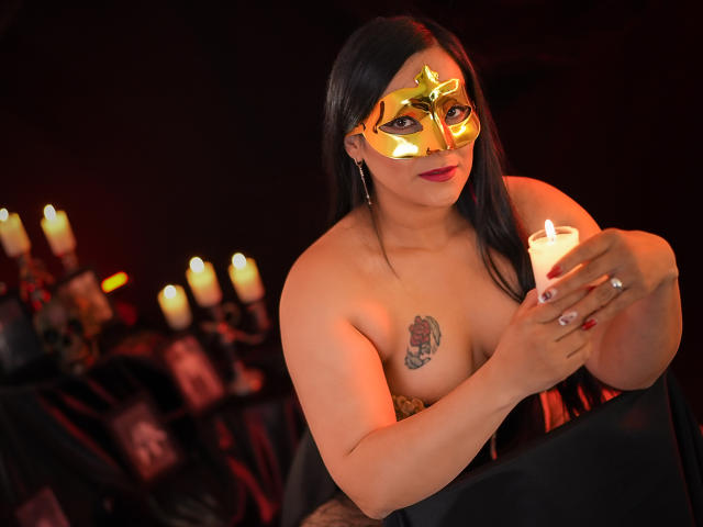 MichellMonet with dark hair, wearing a shiny gold mask, holding a candle, topless, showing her tits, a tattoo on her right boob, red nails, and black background.