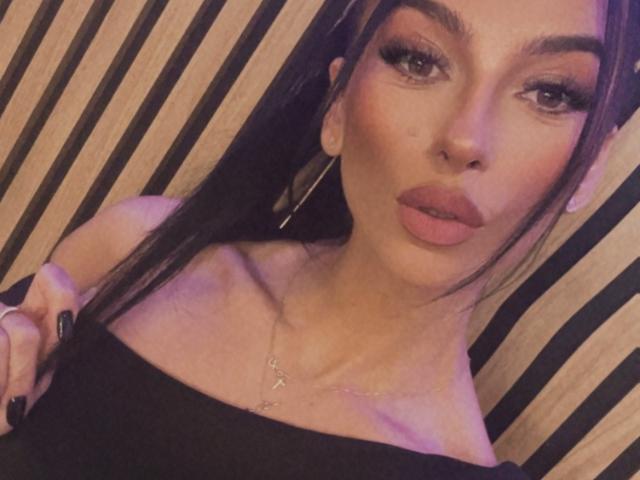 KinkyAmelie with pouty lips, dark long hair, thick eyebrows, long lashes, and hoop earrings, wearing a black off-shoulder top, showing her bare shoulders and collarbone.