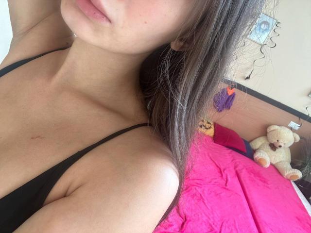 MoonLiza's face and chest in a selfie, showing her black bra and small, slightly visible tits with a small scar on her collarbone. Brown hair, pink bed in background.