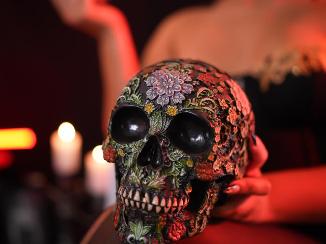 RachellBeaumont's hands hold a skull with colorful floral and vine tattoos, black eyes, and a flower on the forehead. Candles are lit in the background.
