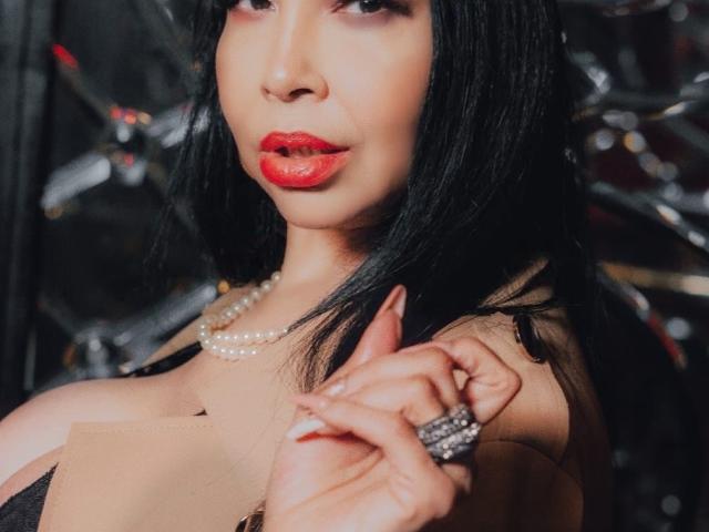 AngelinaDeleon with black hair, red lipstick, pearl necklace, and ring, holding her shoulder, showing her tits in a black dress, looking seductive.