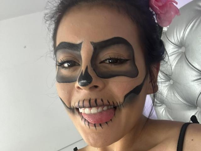 CamilePretty with black face paint resembling a skull, has black eye makeup, nose paint, and black lip lines with a tongue sticking out. She's wearing a pink flower.
