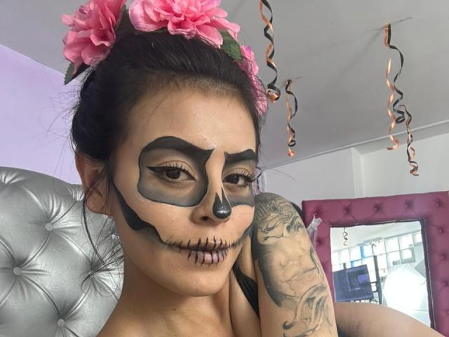 CamilePretty has black face paint with skull eyes and lips, pink flower in hair, tattooed arm, and curly ribbons, sitting on a silver tufted chair, looking at camera.