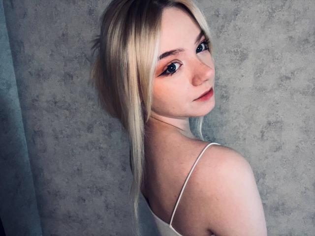 EnchantingEyes has blonde hair, fair skin, and blue eyes. She's looking over her shoulder, wearing a white strap dress, with her bare back and small tits visible.
