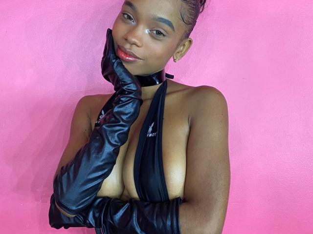 JuicyTitss with dark skin, wearing black leather gloves and a black halter top that exposes her tits, against a pink background, smiling and touching her cheek.