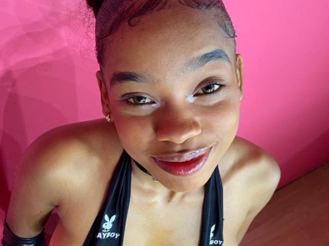 JuicyTitss with shiny lips, black halter top, visible cleavage, and small gold earrings, smiling at the camera, pink background, brown skin, curly hair, and subtle makeup.