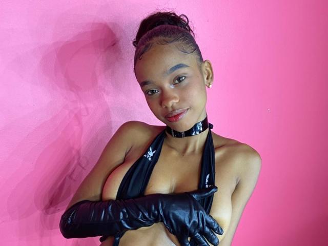 JuicyTitss with dark skin, black glossy gloves, and choker, covering her small tits with arms, wearing a shiny black halter top, against a pink wall.