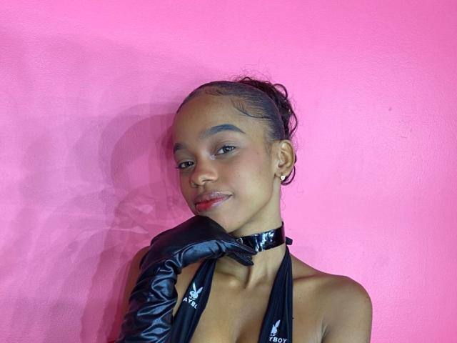 JuicyTitss with dark skin, red lipstick, and black glossy gloves, wearing a black halter top, touching her chin, against a bright pink wall, smiling subtly.