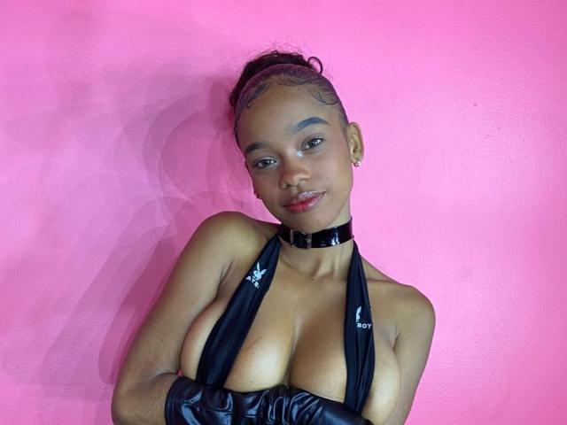 JuicyTitss with dark skin, braided hair, black choker, and black halter top exposing her big boobs, wearing black leather gloves, smiling against a pink wall.