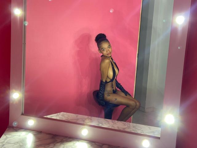 JuicyTitss with dark skin, high bun, black leather strappy outfit, sitting on black stool, reflective mirror, pink wall, illuminated edges, smiling, tattoo on right thigh, sexy.
