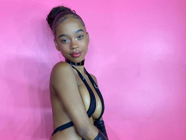 JuicyTitss with dark skin and curly hair in a bun, wearing black strappy lingerie, showing small tits, against a bright pink wall, with a confident, sexy expression.
