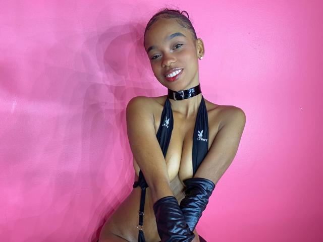 JuicyTitss with dark skin, small tits, wearing black leather choker, halter top, and gloves, smiling against pink wall, looking sexy, showing off her tits and cute face.