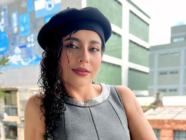 IrinaSweett with curly black hair, wearing a black beret and gray sleeveless top, showing her tits subtly with a confident smile, standing outdoors in sunlight.