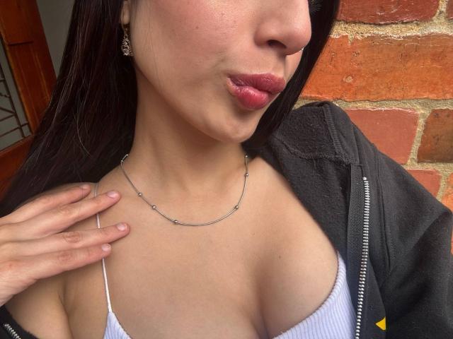 SophieBeaumont with pouty lips, wearing a thin white top showing her tits, black jacket, silver necklace, and earrings, against a brick wall. Her hand touches her chest.