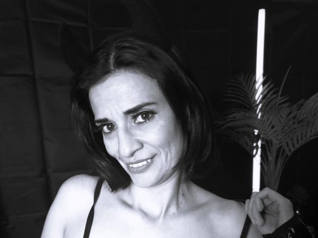 Black-and-white selfie of MaraRoberttss with short dark hair, smiling slightly, showing small breasts, wearing a black strap top, holding a cigarette near her right shoulder.