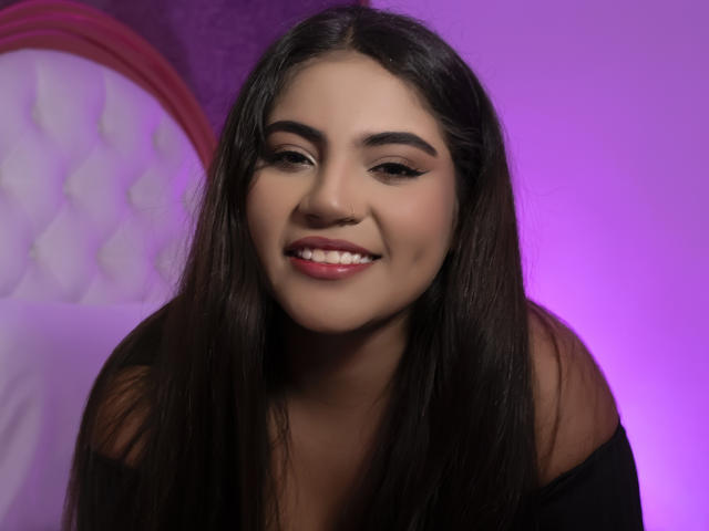 VictoriaRosel with long dark hair, smiling, wearing a black top, showing her ass with a cute smile, pink and purple lighting, and a white tufted bed in the background.