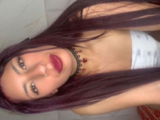 AlisonCold has long purple hair, red lipstick, a nose ring, and a black choker with red beads. She's wearing white pants and looking at the camera.