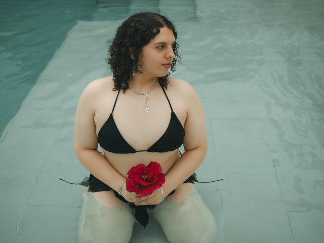 CatleyaNoire mit lockigem, schwarzem Haar, trägt einen schwarzen Bikini, hält eine rote Blume, kniet in einem Pool und zeigt ihre Brüste, während sie ihre Vulva mit den Händen hält..