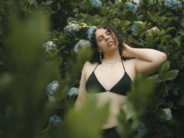 Curvy CatleyaNoire with dark curly hair, black bikini top, and earrings, relaxing in greenery, hand behind head, eyes half-open, looking sexy, lush blue hydrangeas around.